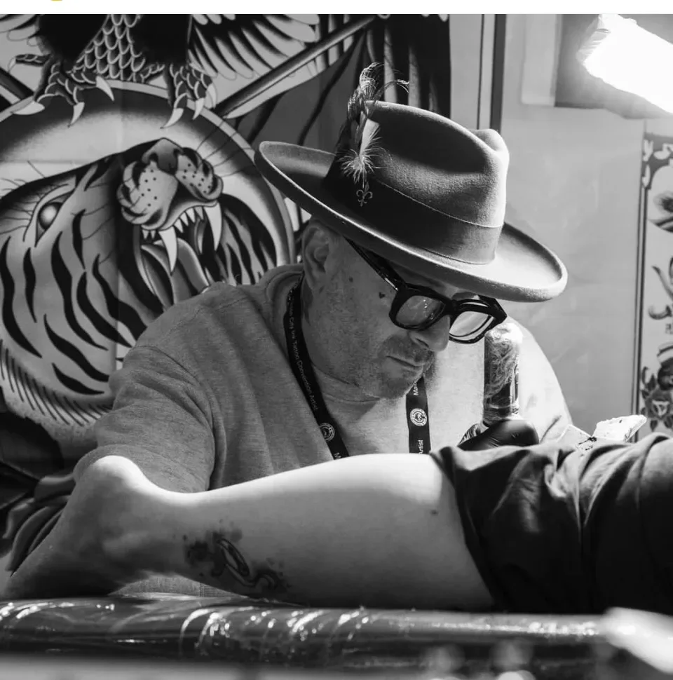 Alessandro Pennella di profilo mentre tatua un braccio, cappello e occhiali.