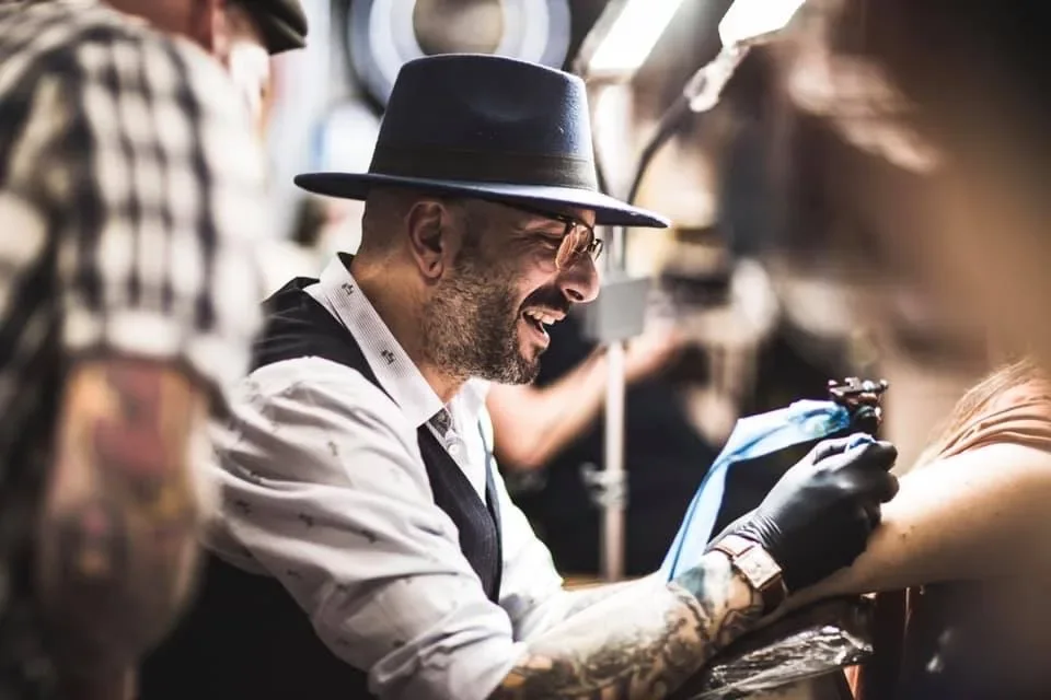 Alessandro Pennella at work in his hat, tattooing an arm and smiling towards the client.