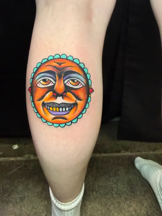 A sun with a human face, orange and teal, on the calf.